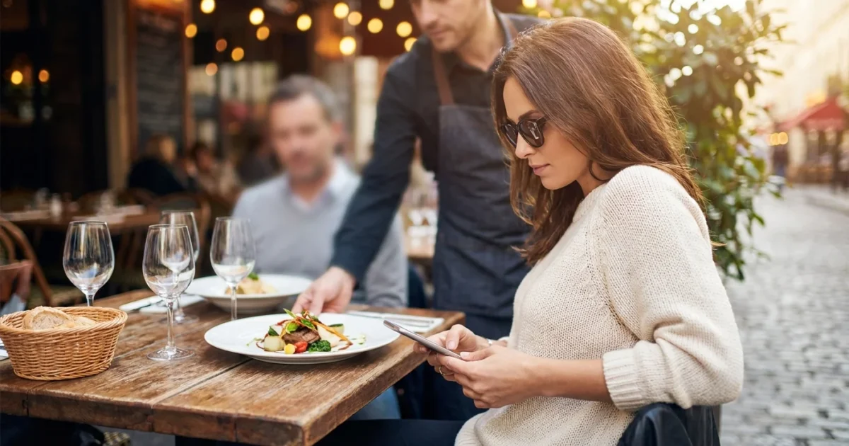 A woman at a restaurant subtly checking her phone while being served a meal.