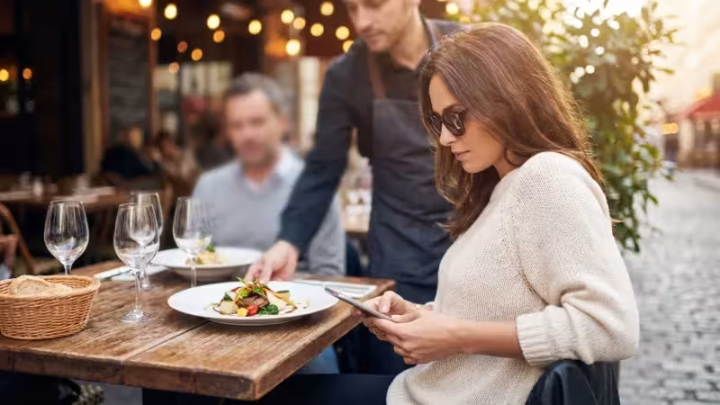 A woman at a restaurant subtly checking her phone while being served a meal.